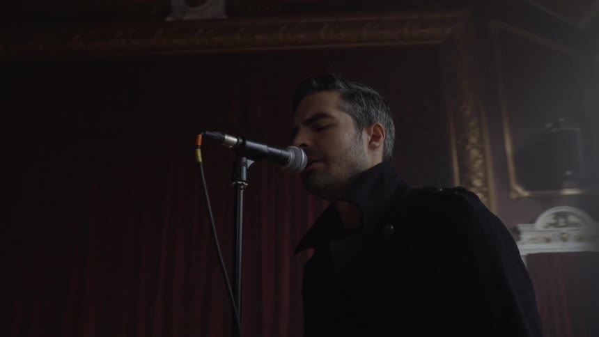Man sings into a microphone while performing with a band in a large room