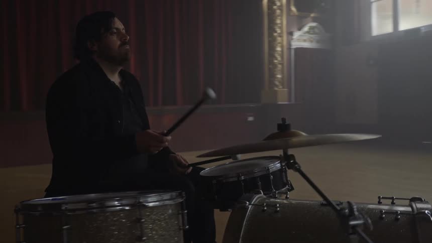 A man plays the drums as the band plays keyboards and sings