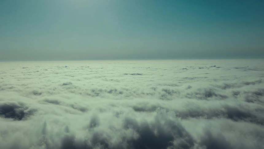An aerial view shows a vast continuous blanket of white clouds stretching to the horizon under a hazy sunlit sky