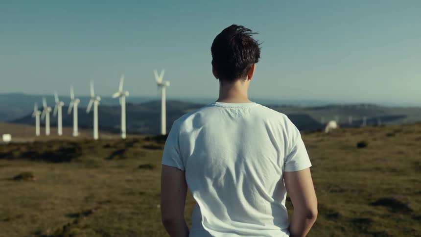 A young man stands on a wind farm on a mostly sunny windy day