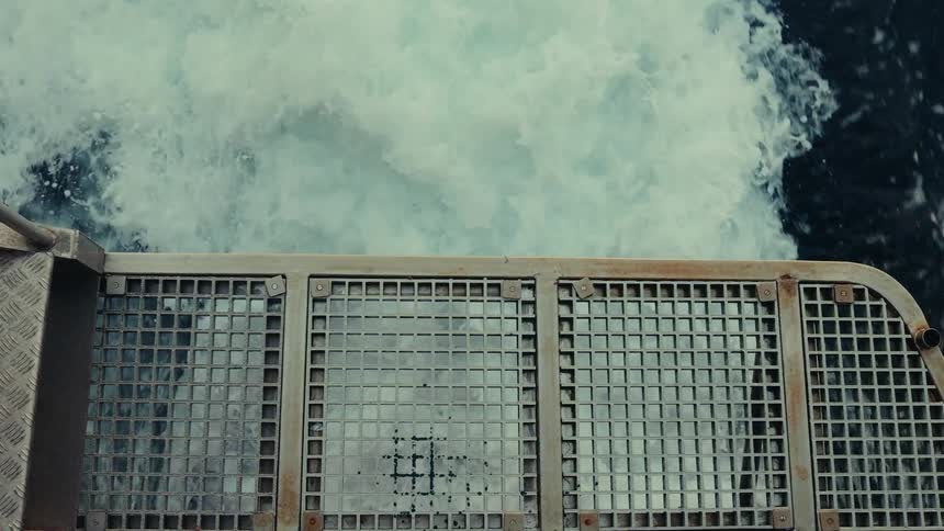 A topdown view from a metal grate platform shows the turbulent white foamy wake of a vessel churning rapidly through darker water