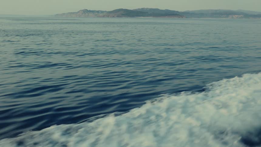 A view from a moving boat captures its white foamy wake against the darker rippling ocean with a hazy distant landmass visible on the horizon