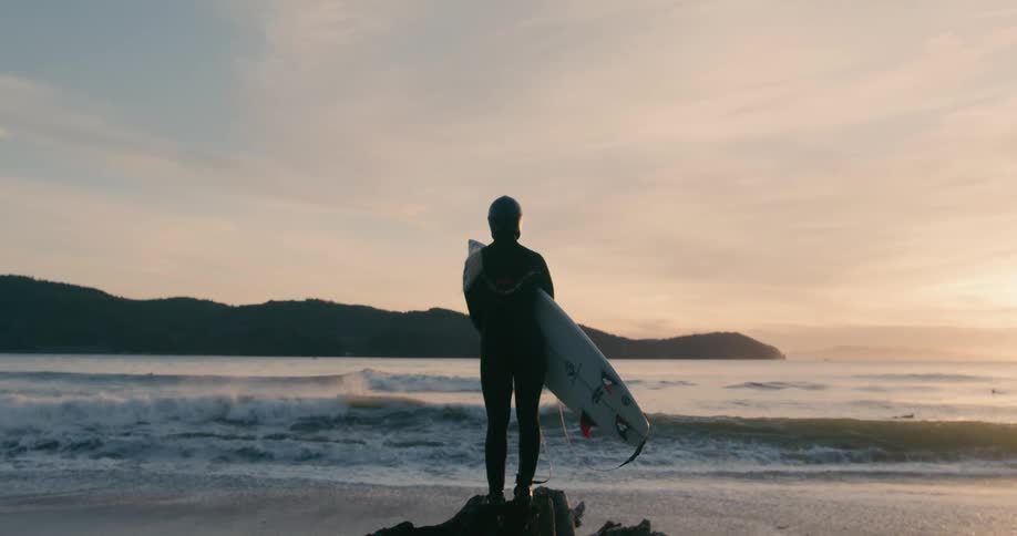 A woman standing on a rock with a surfboard looking at the sea and its waves