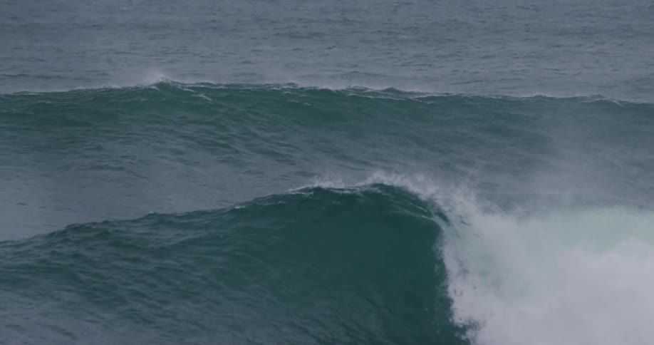 A powerful ocean wave crests and begins to break spraying mist into the air over a choppy sea beneath overcast skies