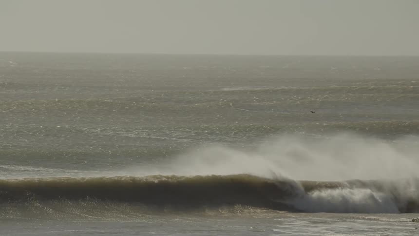 Large powerful ocean waves with significant sea spray blown from their crests break in a rough choppy sea under a hazy sky with birds flying overhead before the view pans to a rocky shoreline