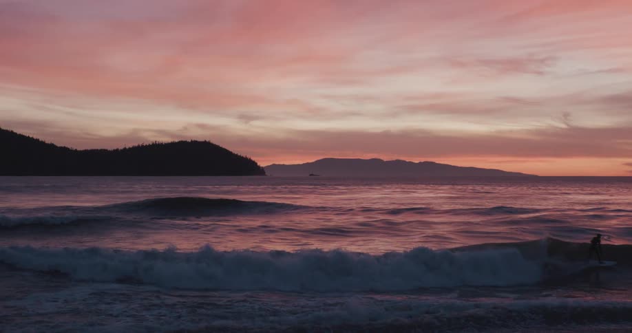 A surfer rides a wave across the ocean under a vibrant pink and orange sunset sky with silhouetted landmasses in the distance after which the scene focuses on the colorful sky reflecting on the rolling waves