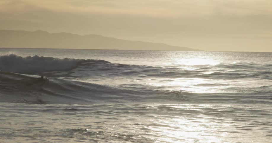 A lone surfer paddles on their board across sunkissed ocean waves towards rolling swells with a hazy distant landmass on the horizon