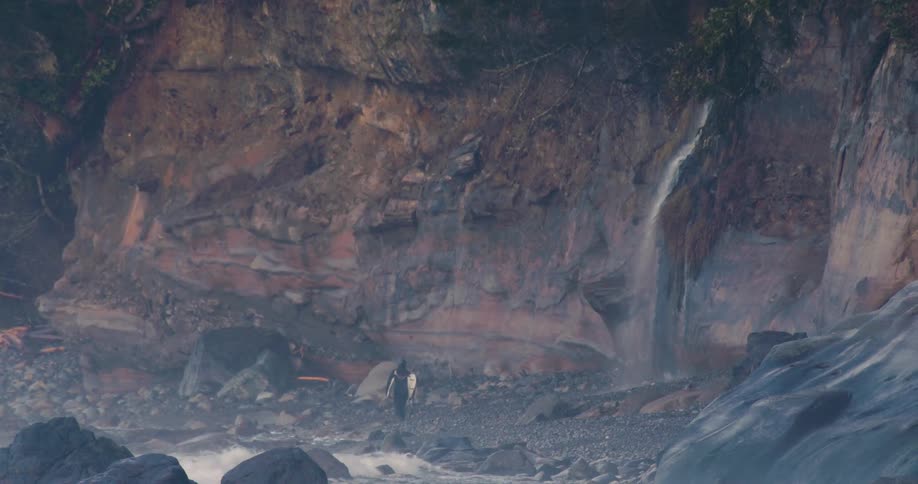 A lone surfer walks along a rocky coastal shoreline at the base of a rugged cliff with a small waterfall trickling down beside them
