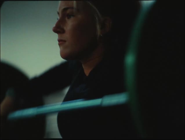 A woman at the gym lifting weights with two men sitting on the floor nearby watching