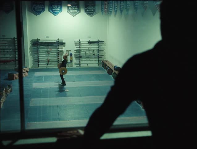 A woman at the gym lifting weights while a man watches from a distance