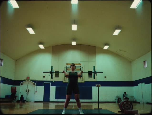 A female weightlifter is captured mid lift in a gymnasium standing on a platform with a barbell raised overhead in a powerful pose