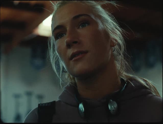 A woman with blonde hair is indoors wearing a red hoodie and headphones around her neck