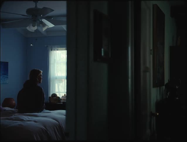 A woman sitting on a bed looking sad as a younger woman appears at the door watches for a moment and then walks away