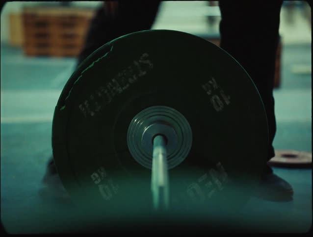 A closeup shot captures a green weight plate on a barbell as hands adjust it with slightly blurred gym equipment in the background