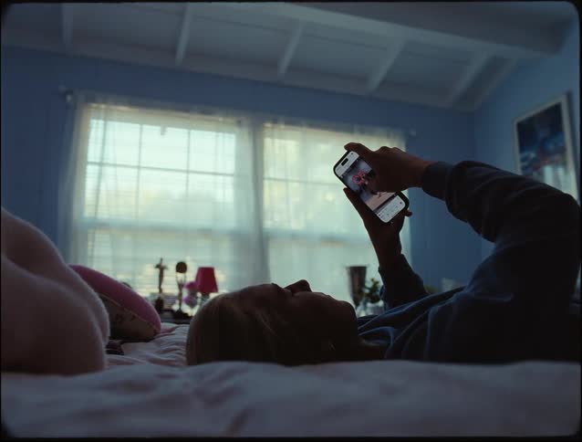 A woman lying on a bed in her room scrolling through social media on her phone