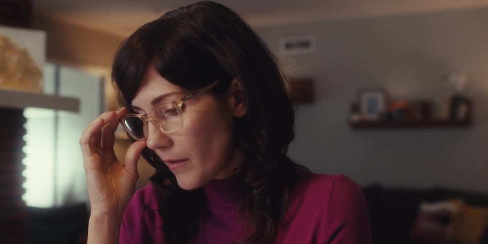 A woman wearing a red dress is indoors talking and removes her glasses during the conversation at midday