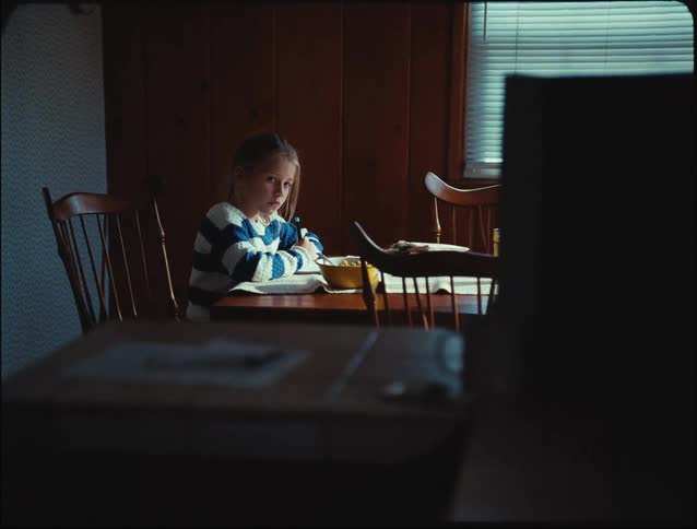 A young girl sitting in the dining doing her home work looking sad with a plate of food beside her as her mother walks by