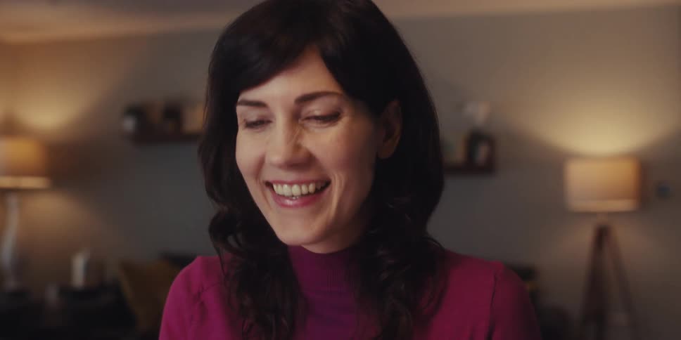 A woman smiles and looks downward while speaking appearing to enjoy a video call in a warmly lit room