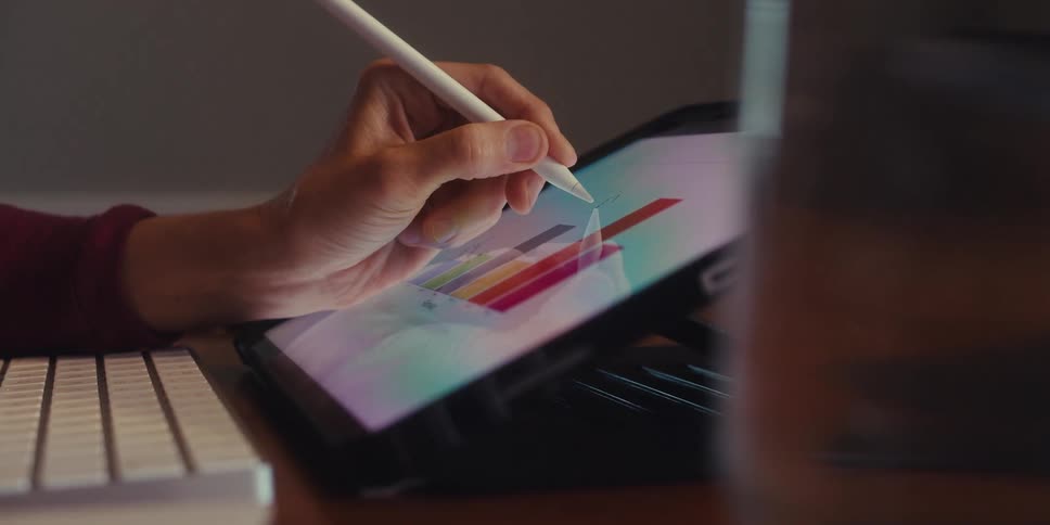 A hand uses a white stylus to interact with a colorful bar graph on a tablet screen analyzing data with a keyboard and mug nearby