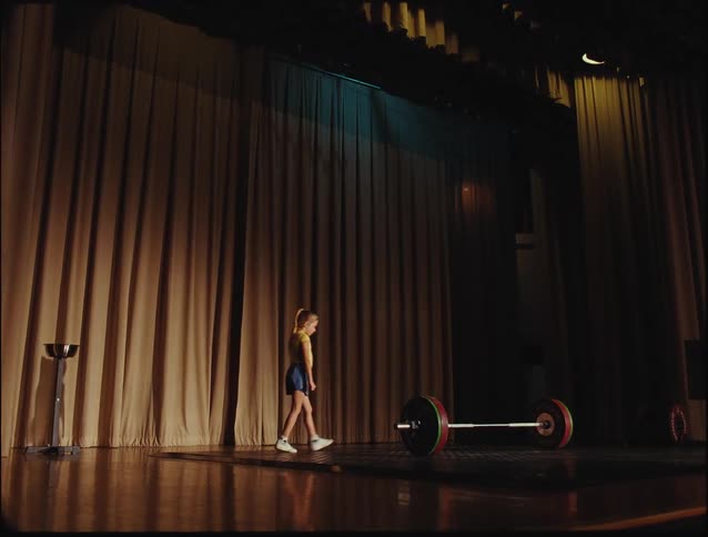 A child walks up a stage and tries to lift a heavy weight