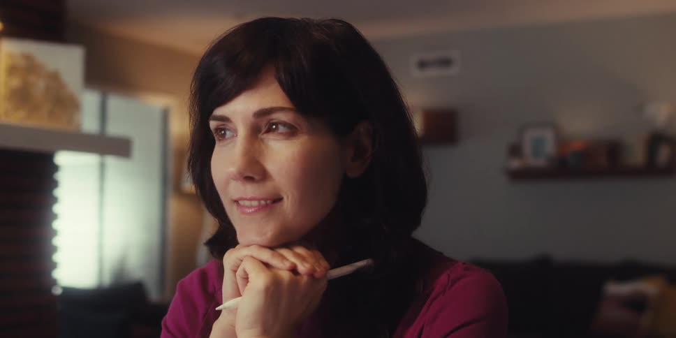 A woman sits indoors thoughtfully gazing off screen with her hands clasped under her chin holding a pen