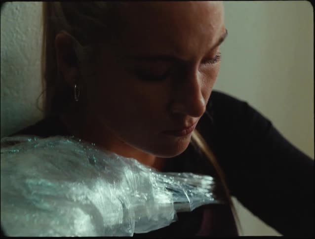 A woman sits looking sad with one hand resting on her forehead and several layers of plastic cellophane wrapped around her shoulders