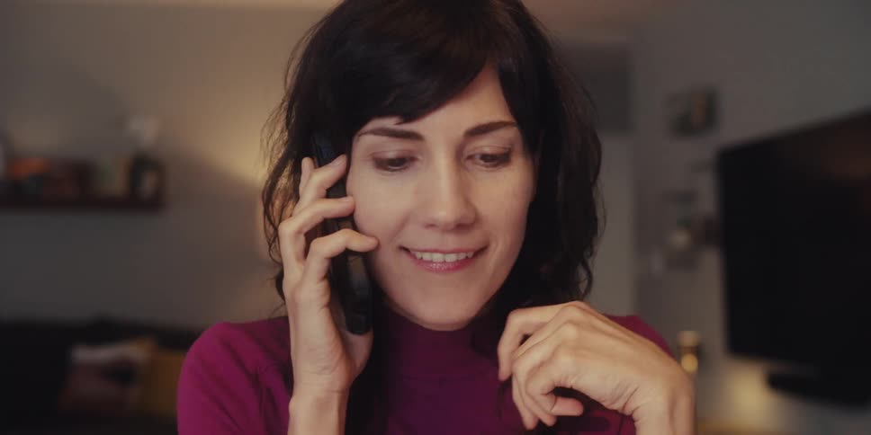 A woman in a living room talking on the phone and smiling then drinking water