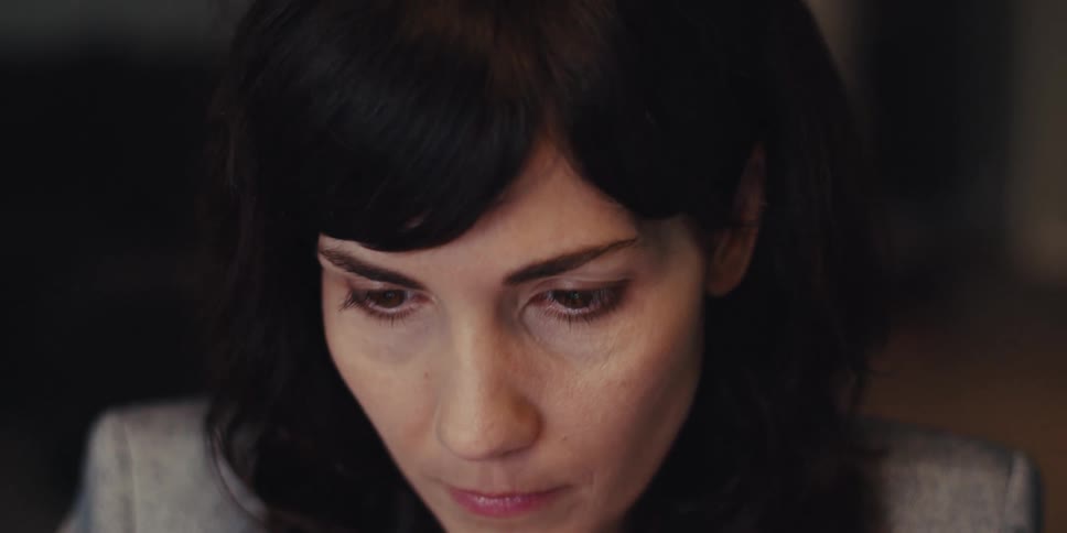 A woman with long black hair wearing an ashcolored suit is typing on a keyboard and looks tired