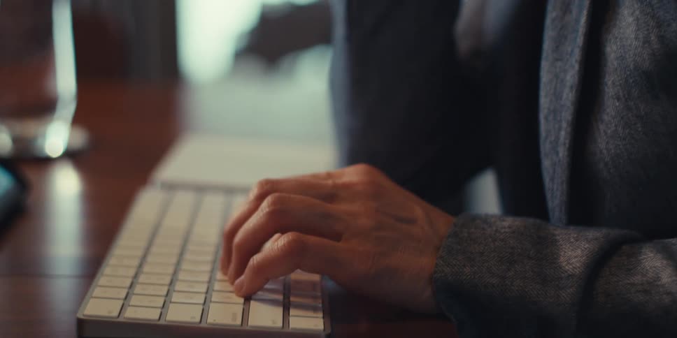 A young woman with long black hair is in her office wearing glasses and a blazer suit while typing on a keyboard