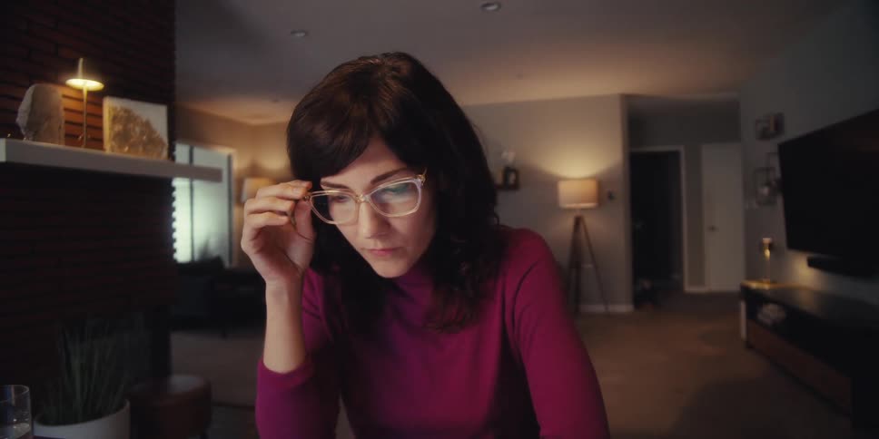 A woman wearing a red dress walks up to a desk in her living room sits on a chair puts on glasses picks up a pen and writes then picks up her phone to make a call while laughing during the conversation