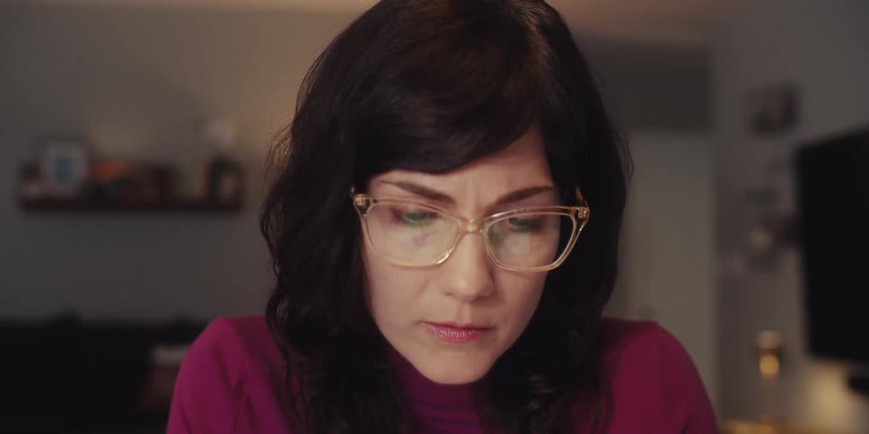 A young woman wearing glasses sits in front of her computer smiling and laughing as she speaks during a conference call