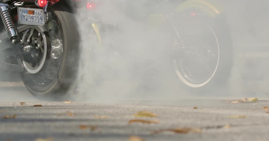 Two people rev motorcycle engine in park to make back tires smoke