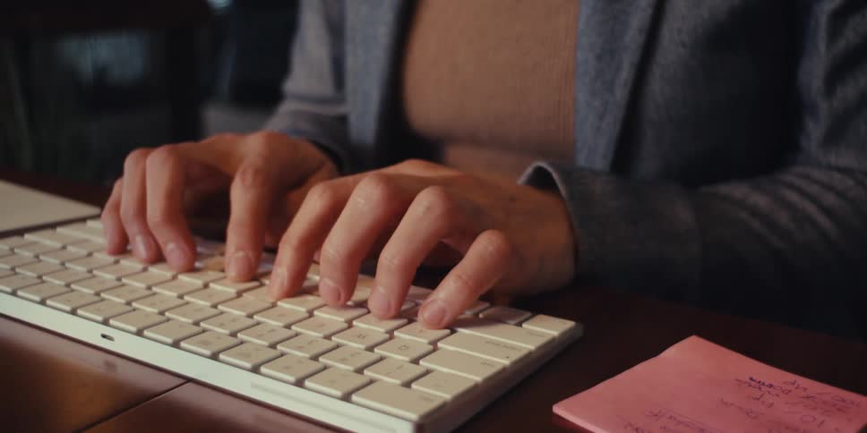 A young woman in her office is typing on her keyboard and laughing while working on her computer wearing a blazer suit and glasses
