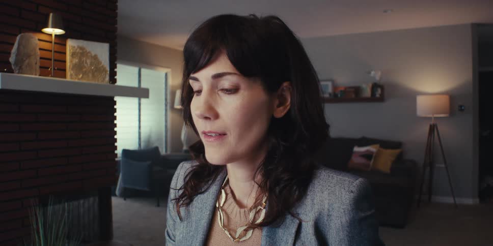A woman in professional attire appears to be engaged in a video call or moment of introspection in a modern warmly lit living room
