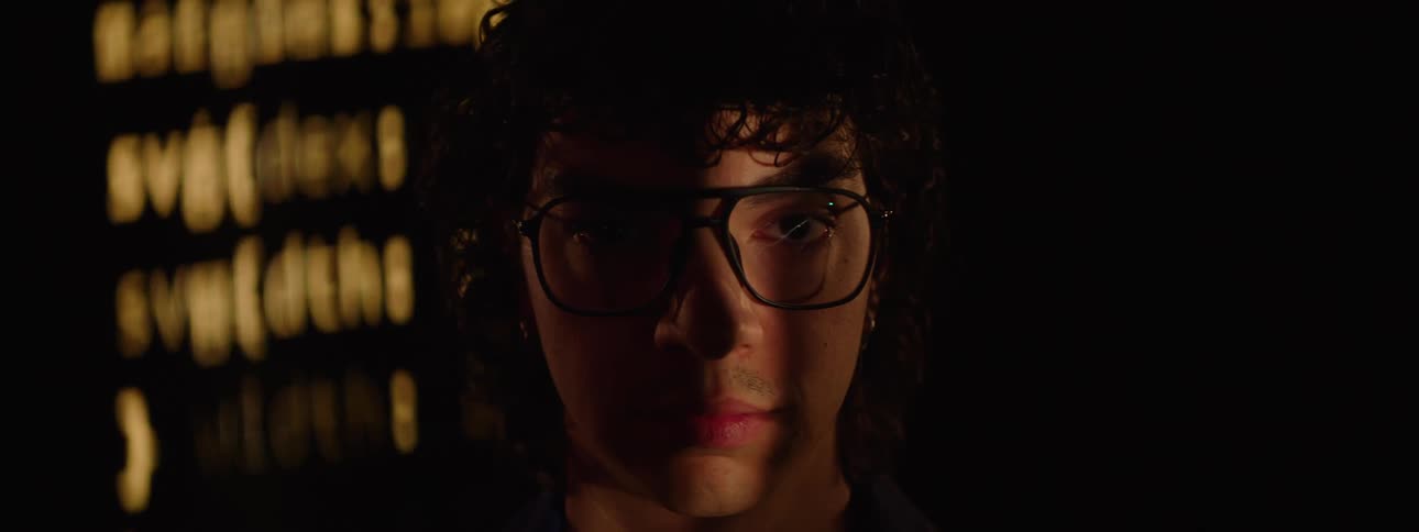 A person wearing glasses with part of their face in shadow against a dark background with blurry yellow text