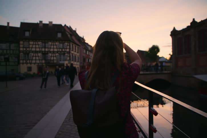 A woman with a backpack snaps a photo at sunset by a canal in a charming european town