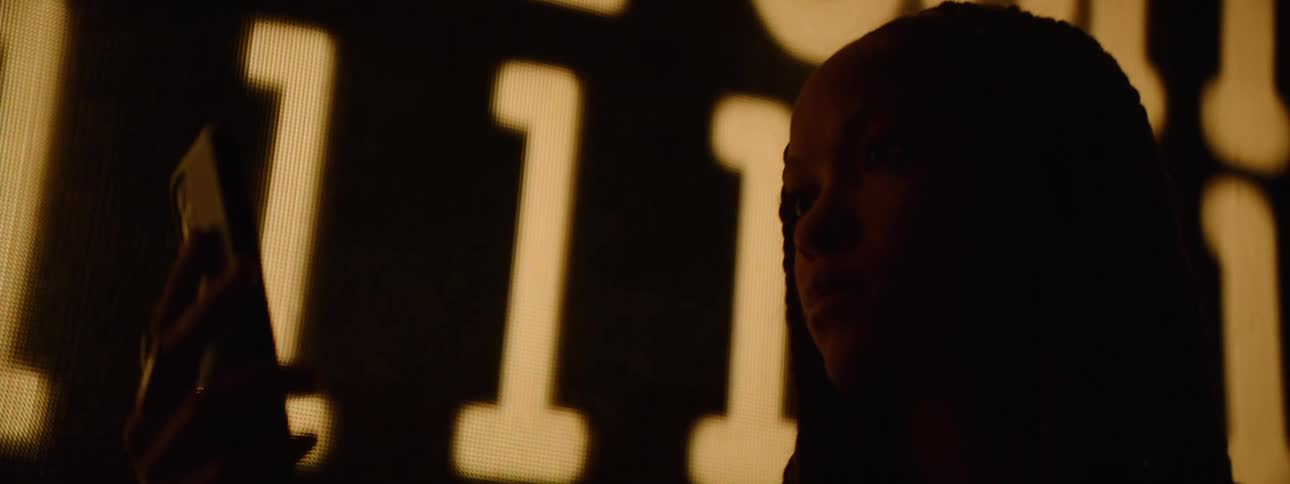 The face of a woman who is passing by very dark and she is using her cell phone and behind her is a sign with yellow lights