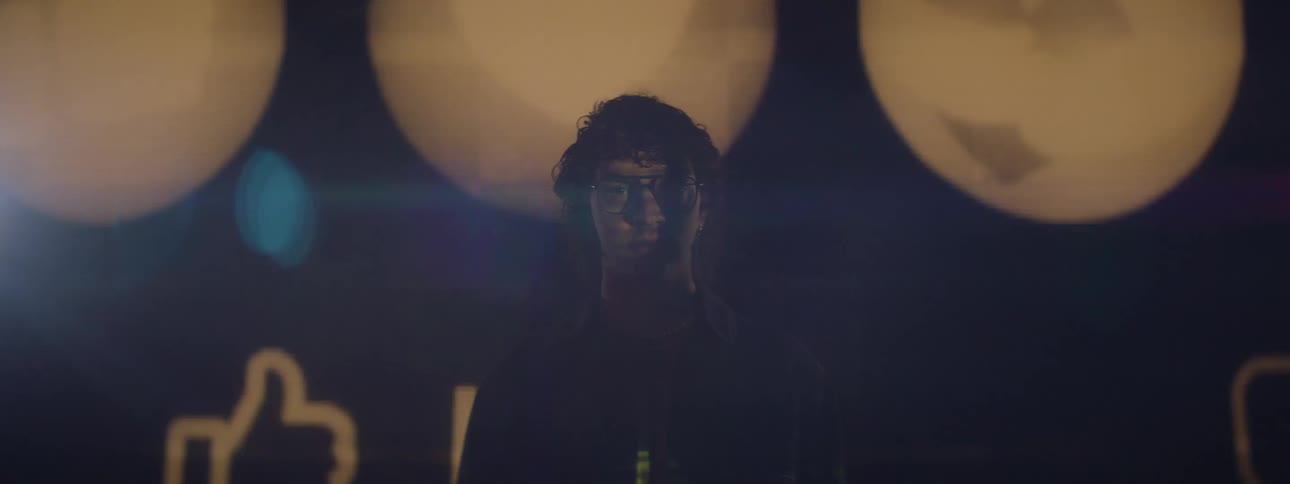 The face of a young person wearing glasses who is standing in front of a huge screen with yellow lights