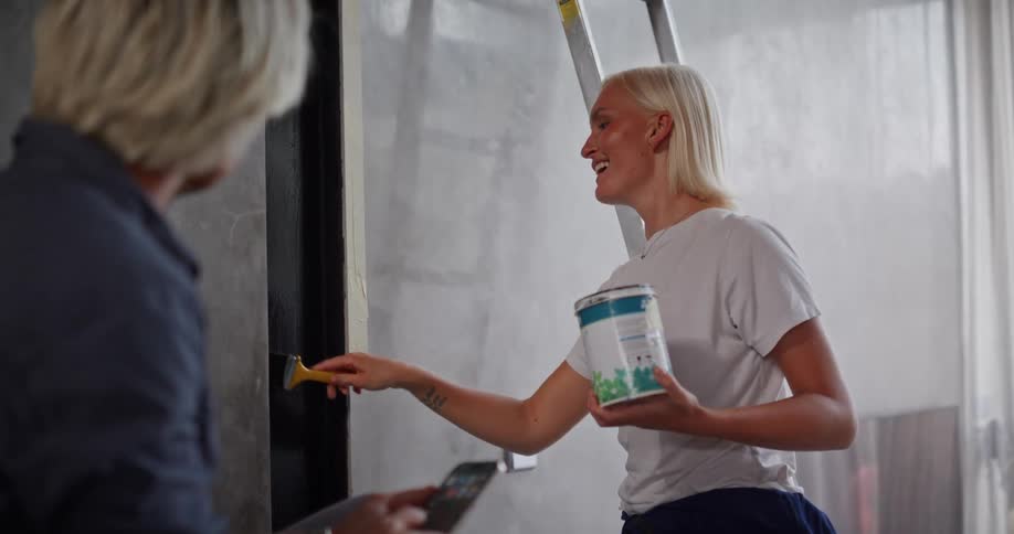 A woman smiles while painting a wall and talking with another person showing a moment of teamwork and casual conversation during a home project