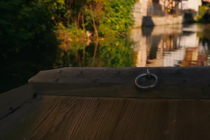 The edge of a wooden boat with a metal ring with a canal reflecting surrounding buildings and lush vegetation in the background