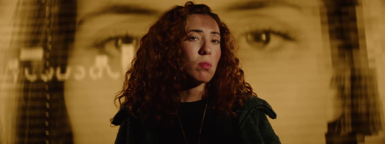Portrait of a woman with curly hair and in the background a screen displaying faces