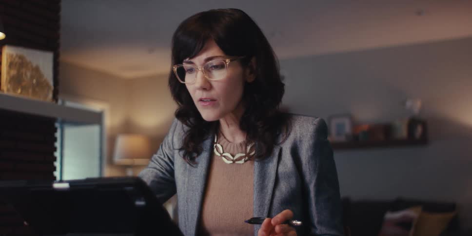 A woman in a blazer puts on her glasses and interacts with a tablet appearing engaged in a workrelated video call from her home