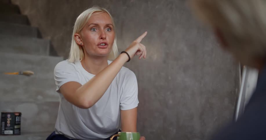 A woman is talking to a man about painting while sitting on the stairs and holding a coffee cup