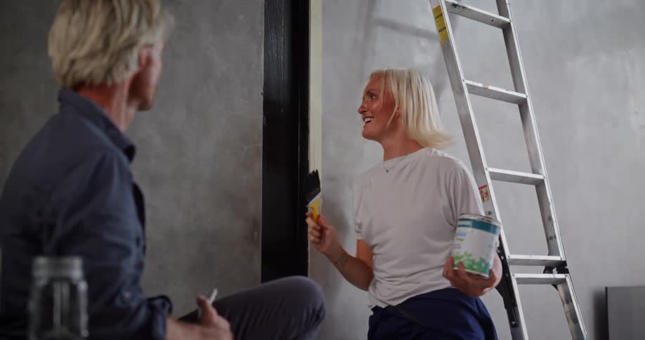 A woman is smiling and dancing playfully while painting a wall holding a paint can in one hand and a brush in the other