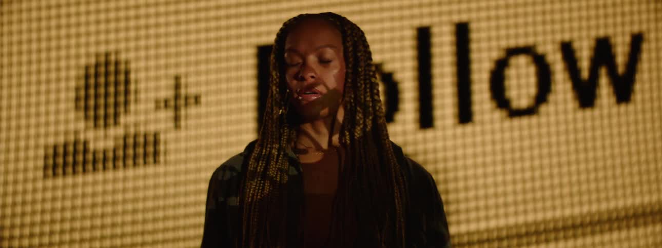 A woman with long braided hair holds a smartphone while standing in front of a large screen displaying a bold social media follow button