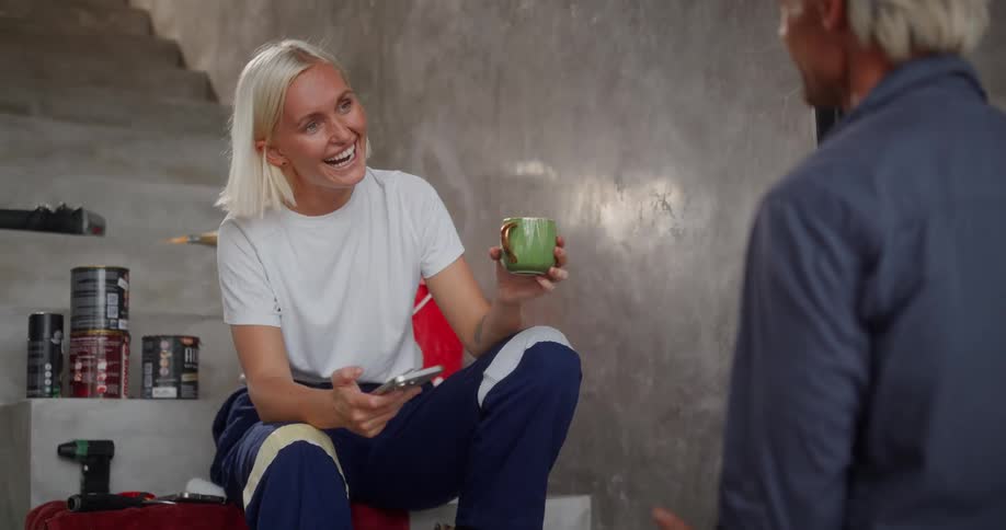 Woman sitting on stairs laughing while holding a smartphone and a mug and talking to a man