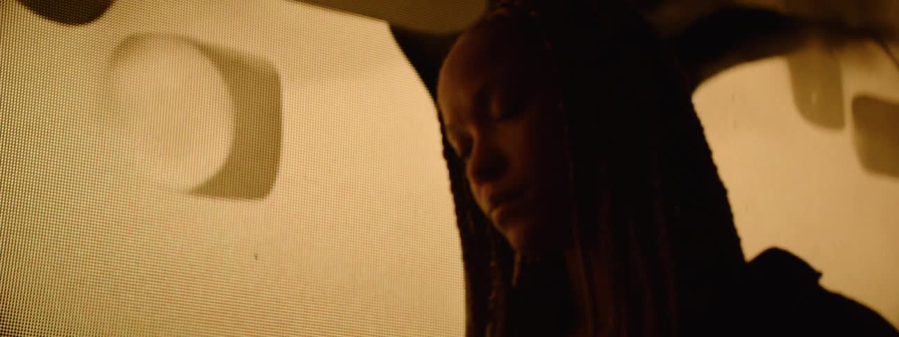 A young woman with braids looks at her phone in low moody lighting appearing thoughtful
