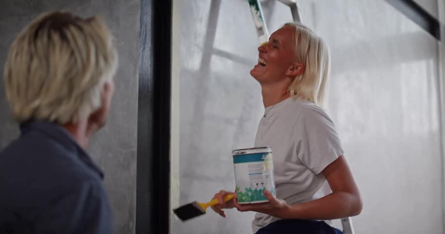 A blondehaired woman paints a dark trim on a lightcolored wall while conversing with a man beside her