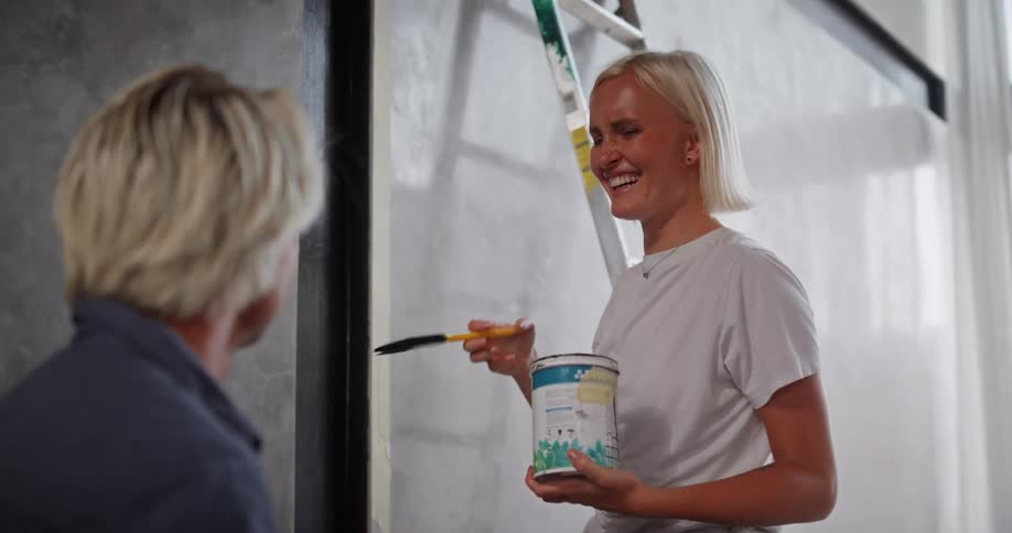 A woman holding a paint can and brush is smiling while conversing with a man during a home painting project
