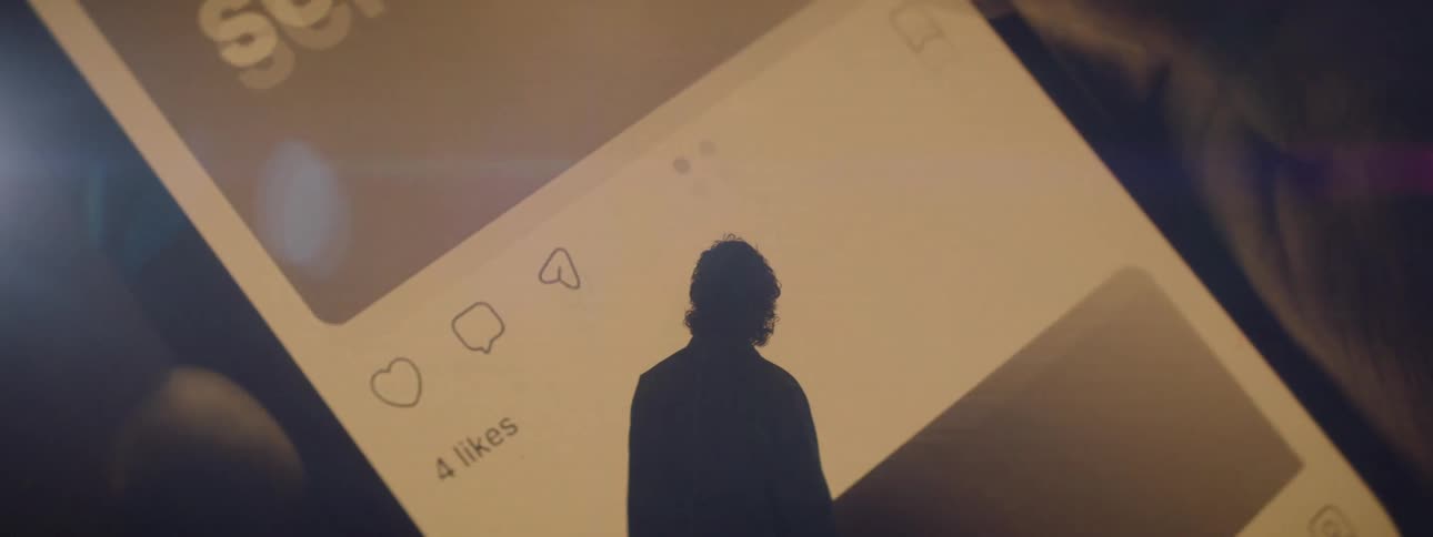 Man looking at a large screen displaying a cell phone on a social media app then looking at his own phone